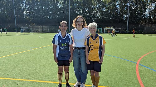 Alison Bennett MP stands between two Burgess Hill Hockey Club players at the club’s season kick-off event. The three are smiling on the hockey pitch, with players and matches visible in the background