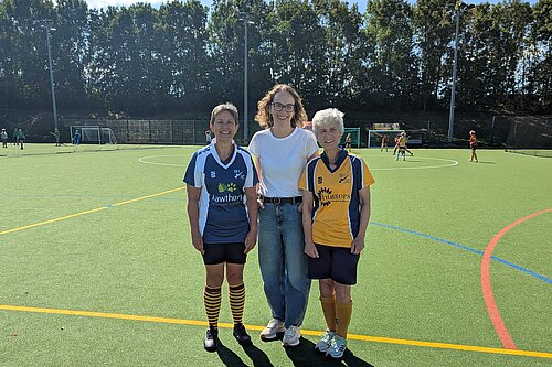 Alison Bennett MP stands between two Burgess Hill Hockey Club players at the club’s season kick-off event. The three are smiling on the hockey pitch, with players and matches visible in the background