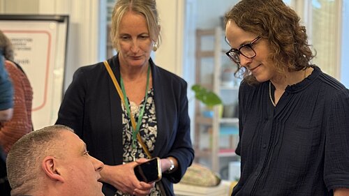 Alison Bennett MP speaks with attendees at a carers’ event, standing beside a seated man and another woman in a bright community room.