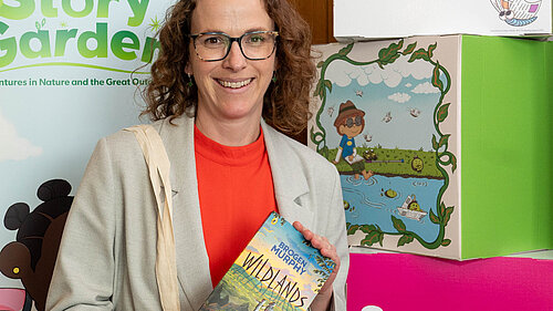 Alison holding the book 'Wildlands' by Brogen Murphy in front of banners promoting the The Reading Agency’s 2025 Summer Reading Challenge.