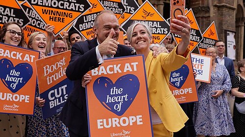 Ed Davey and Daisy Cooper take a selfie at a Liberal Democrat event, holding orange “Back at the heart of Europe” placards. Alison Bennett MP is visible at the far left, alongside supporters holding Liberal Democrat and “Winning Here” signs.