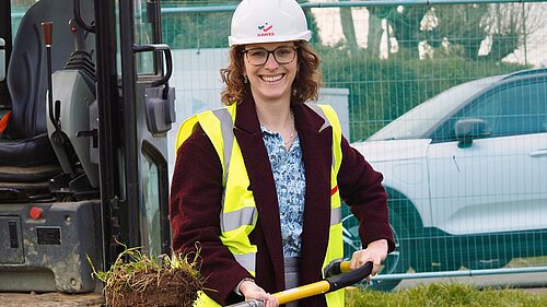 Alison Bennett MP holding a spade with a turf next to a digger near haras fencing