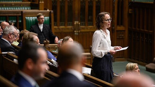 Alison Bennett MP in standing and speaking parliament chamber with speakers chair in background and other MPs around
