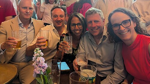 Alison and other Liberal Democrat MPs in a pub watching the England Vs DRC football match, seated around a small wooden table, and other people behind.