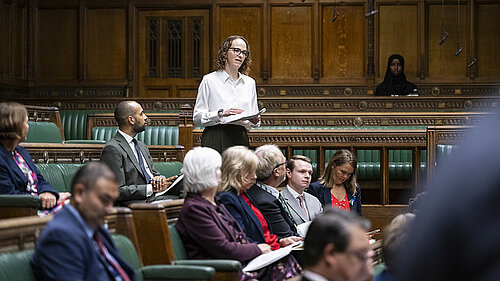 Alison in Parliament Chamber asking a question while standing, with other MPs sitting around